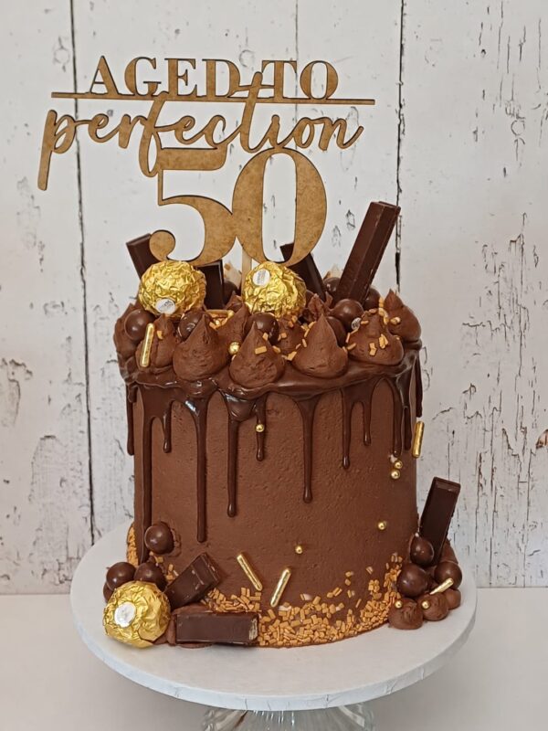 IMG-20261003-WA0027 Chocolate drip cake with piped chocolate swirls, assorted chocolates, gold sprinkles and a gold “Aged to perfection 50” topper.