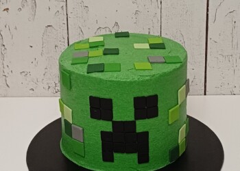 Round green Minecraft Creeper cake with a black pixel face, green square decorations and Xander lettering on the cake board.