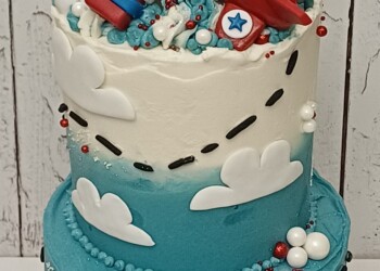 Two-tier blue first birthday cake with a red aeroplane topper, white clouds and a dotted flight path.