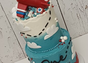 Angled view of a two-tier aeroplane birthday cake with blue frosting, white clouds, red trim and a flight-path design.